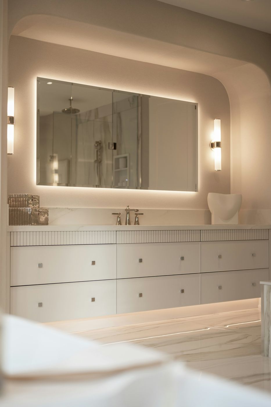Bathroom vanity with a large backlit LED mirror, two fluted glass wall sconces and toe-kick LED lighting under a floating vanity