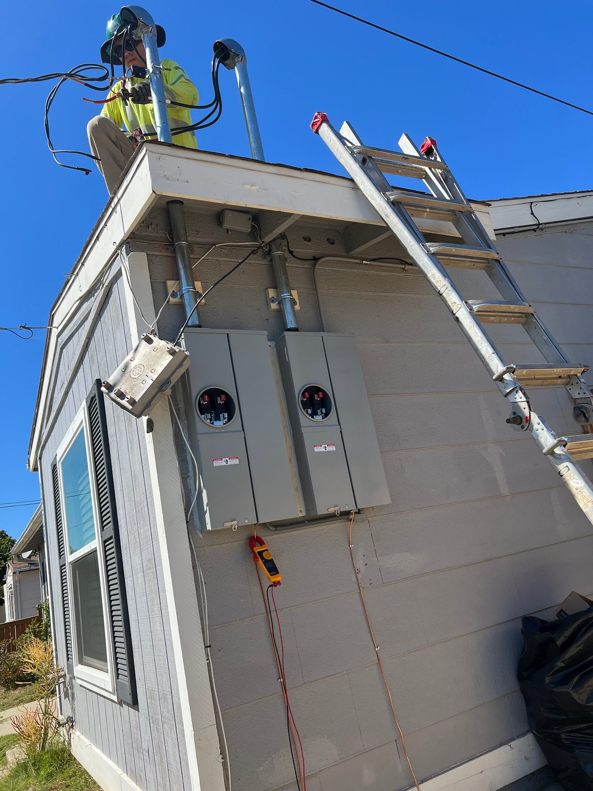 A small house with two new meter sockets on the wall, a ladder against the roof and an electrician working at the service mast