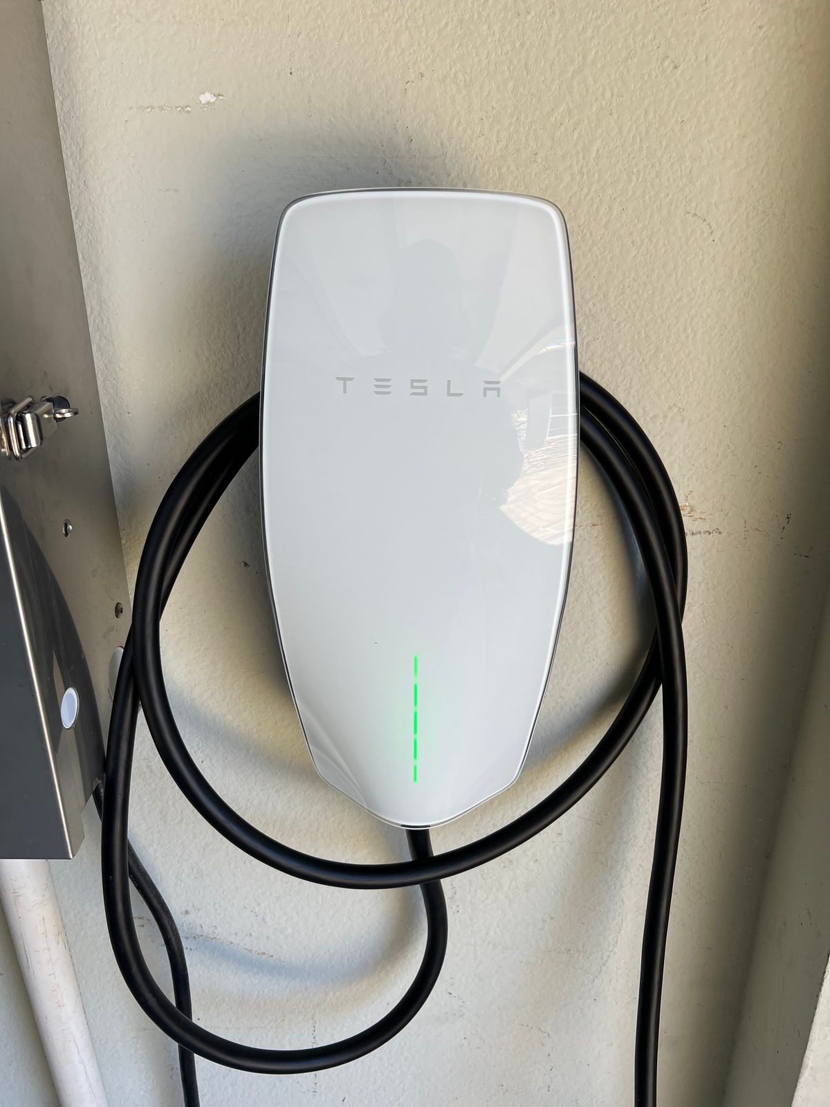 White Tesla Wall Connector mounted on a wall with its charging cable coiled around it and the green status light on